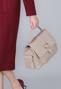 Borsa beige con un design strutturato, grande chiusura a ribalta, dettagli in oro e manico superiore. Il materiale sembra essere liscio e sintetico.