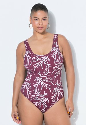 Femme aux cheveux courts portant des boucles d'oreilles créoles dorées et un maillot de bain une pièce violet avec des motifs de corail blancs, se tenant devant un fond blanc.