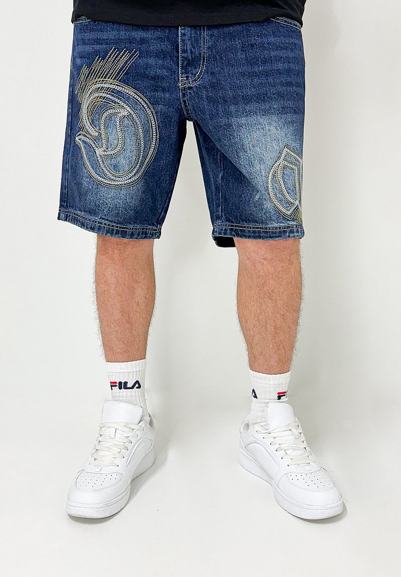 Denim shorts in a dark wash, featuring decorative embroidery in grey and gold. Worn with white sneakers and logo socks.