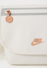 Borsa a tracolla bianca in tessuto con una texture liscia, zip grigia e logo Nike arancione. Presenta una chiusura a pattina e una tracolla regolabile.