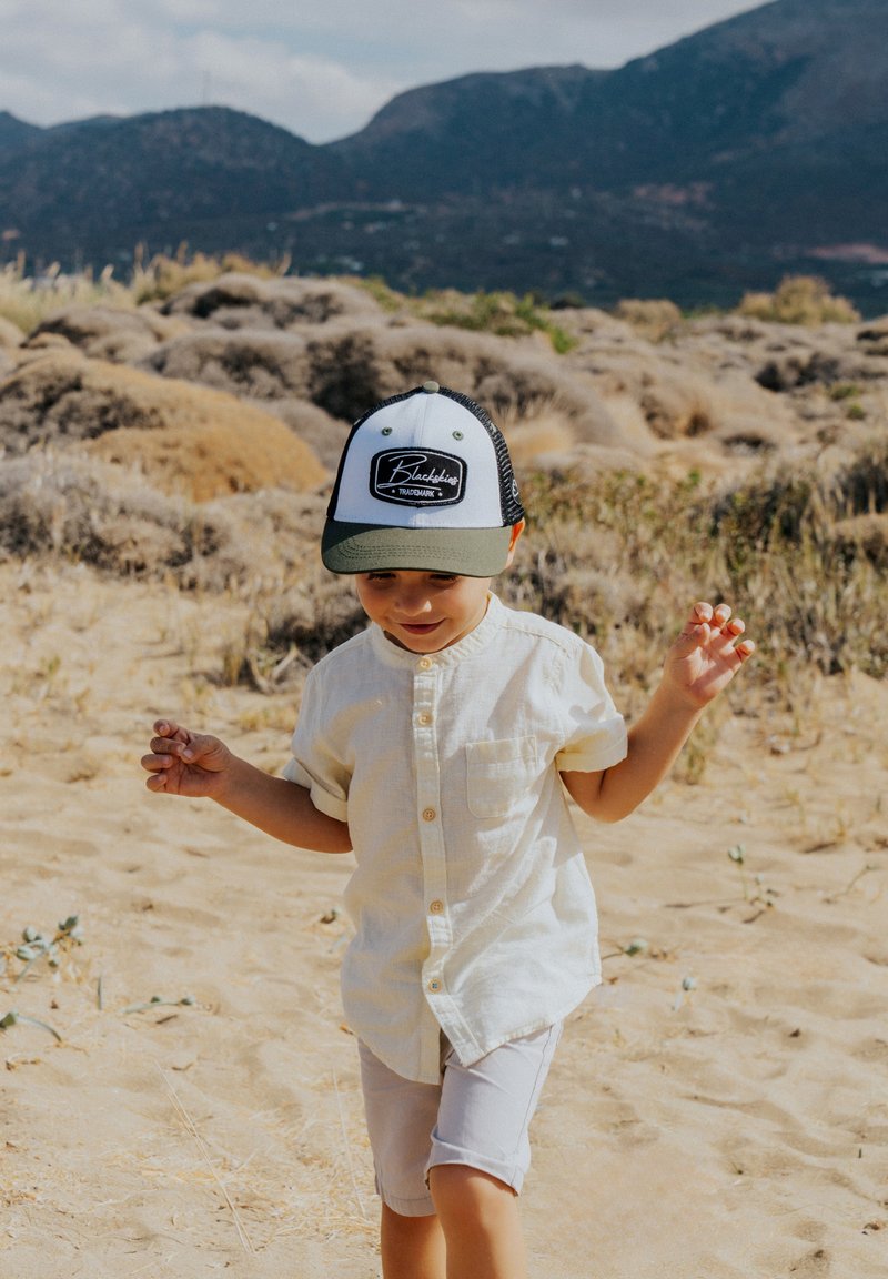 Niño vestido con una camiseta de manga corta amarilla clara y pantalones cortos beige, con una gorra en blanco y negro que presenta un logo, caminando por un terreno arenoso.