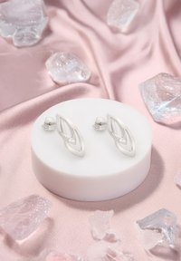 Silverfärgade örhängen i bladform med polerad yta och en rad klara stenar. Visas på en rund vit pedistal mot en bakgrund av rosa tyg.