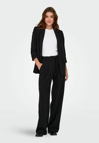 Donna in piedi con una mano in tasca, che indossa un blazer nero, pantaloni ampi neri con cintura, camicia bianca e scarpe nere su sfondo bianco.