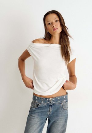 Junge Frau mit langen braunen Haaren, die ein schulterfreies weißes Oberteil und hoch taillierte blaue Jeans mit Metallknöpfen trägt.