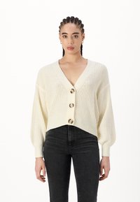 Crèmekleurige gebreide cardigan met een diepe V-hals, oversized mouwen en vier grote, ronde knopen aan de voorkant. Gepaard met zwarte jeans.