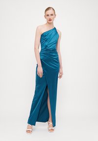 Saten, enostrani teal večerna obleka z draperijami in razporkom do stegna. Model nosi bele sandale na peto.