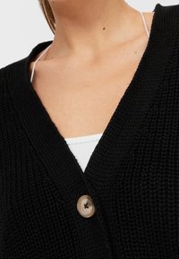 Gros plan sur une personne portant un cardigan noir en maille avec un grand bouton unique au-dessus d'un haut côtelé blanc à fines bretelles.