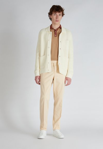 Cardigan crema a maglia con texture a costine, polo beige e pantaloni beige chiaro; modello che indossa scarpe bianche, in piedi su uno sfondo neutro.