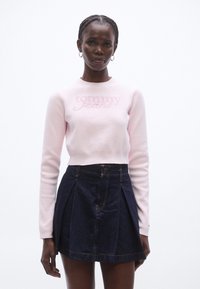 Maglione rosa chiaro corto con la scritta "Tommy Jeans" sul davanti, abbinato a una gonna plissettata in denim scuro con una tasca frontale e cuciture dorate.