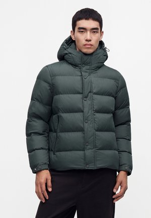 Mann trägt eine dunkelgrüne gesteppte Winterjacke mit hohem Kragen, Kapuze, vorderen Druckknöpfen und Seitentaschen vor einfarbigem Hintergrund.