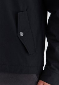 Crna jakna s džepom na preklop zatvorenim dugmetom, teksturiranom tkaninom i elegantnim, minimalističkim dizajnom. Vidljivo šivanje duž donjeg ruba.