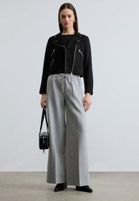 Γυναίκα που στέκεται, φοράει μαύρο κομμένο σακάκι και φαρδιά γκρι παντελόνια, κρατώντας μαύρη τσάντα χειρός με λουριά ώμου σε απλό φόντο.