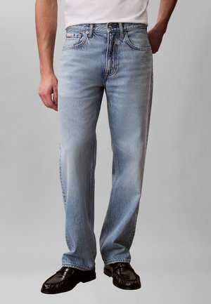 Άνδρας που φοράει ανοιχτό μπλε τζιν ίσιας γραμμής, άσπρο πουκάμισο μπουρνουζομένα μέσα στο παντελόνι και μαύρα δερμάτινα loafers, στέκεται μπροστά σε γκρι φόντο.