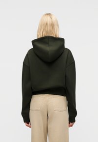 Ξανθιά γυναίκα με την πλάτη γυρισμένη, φορώντας σκούρο πράσινο κοντό φούτερ με κουκούλα και μπεζ παντελόνι ψηλόμεσο με πίσω τσέπες, μπροστά σε απλό φόντο.