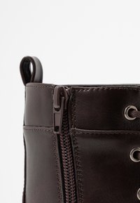 Bottine en cuir marron avec une fermeture éclair latérale, des œillets pour les lacets et des détails de couture. Texture lisse avec une petite languette en haut.