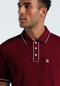 Polo bordeaux avec des rayures blanches contrastantes sur le col et la patte de boutonnage, comportant une poche et un petit logo sur la poitrine.