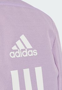 Lila texturerad tyg med en vit Adidas-logotyp och tre vertikala ränder. Designen inkluderar ett subtilt vävmönster.