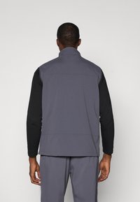 Gray sleeveless vest with a high collar, textured fabric, and contrasting black long sleeves beneath. Simple design with no visible hardware.