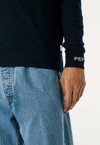Suéter de punto navy con puños y dobladillo acanalados, que presenta la inscripción "PEP" en blanco. Combinado con jeans de mezclilla azul claro.
