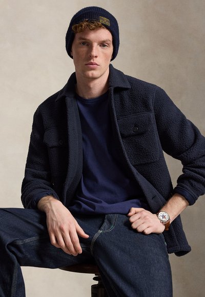 Νέος άνδρας καθισμένος, φοράει ναυτικό μπλε φλις μπουφάν, ναυτικό μπλε σκουφί, σκούρο τζιν, ναυτικό μπλε πουκάμισο και καφέ ρολόι χειρός με δερμάτινο λουράκι, μπροστά σε ουδέτερο φόντο.
