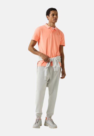 Muž v broskvové polokošili a světle šedých teplácích s odpovídající mikinou uvázanou v pase, stojící na bílém pozadí.