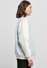 Svetlomodrá univerzitná bunda s krémovými rukávmi, s rebrovanými manžetami a lemom. Obsahuje bočnú vrecko a minimalistický dizajn.