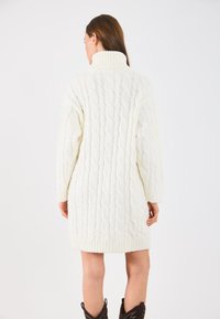 Vestido de punto trenzado en crema, con un cuello alto, dobladillo acanalado y mangas largas con un patrón de punto texturizado.