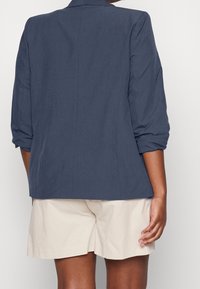 Syy navy blue, kevyt bleiseri, jossa on kolmennesosa hihat ja rento istuvuus. Silkkinen pinta ja hienovaraiset tikkausdetailit. Yhdistetty vaaleisiin shortseihin.