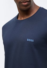 Homme portant un t-shirt à col rond bleu marine avec le logo "BOSS" brodé en bleu clair sur la poitrine, recadré au niveau du cou et des épaules.