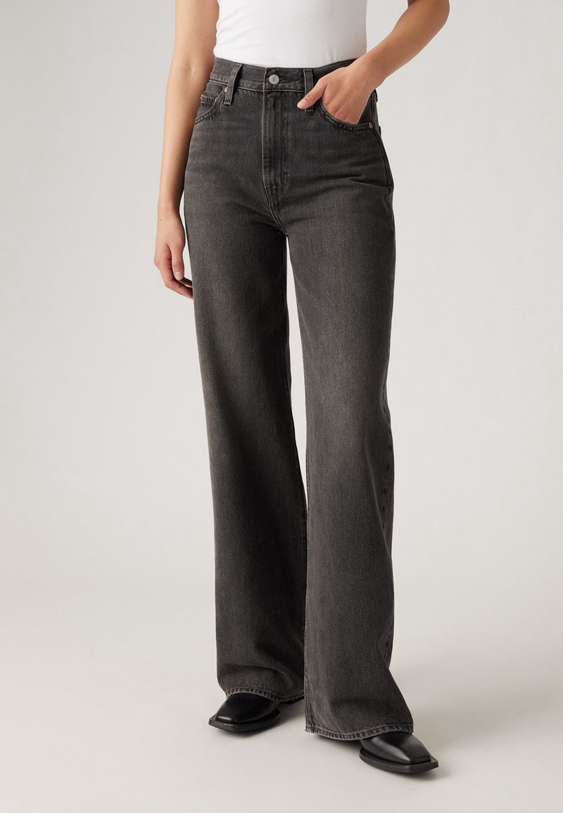 Personne portant un jean noir taille haute à jambes larges avec cinq poches, associé à un haut blanc sans manches et des chaussures noires à bouts pointus.