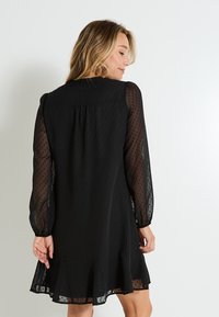 Vestido negro con mangas largas de tul abullonadas y transparentes. Presenta una textura punteada y un dobladillo acampanado. La parte trasera tiene gathers suaves y un escote redondo.