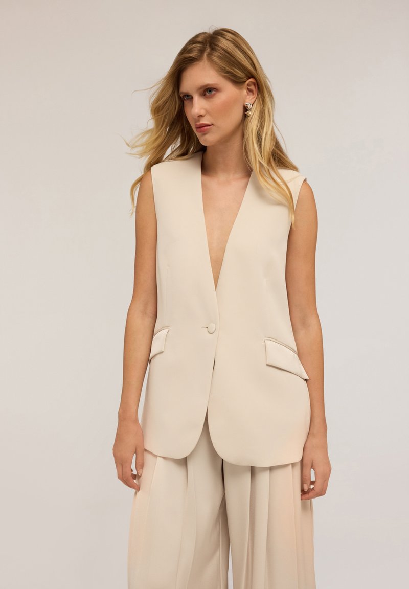 Beige mouwloze blazer met een diepe V-hals, enkele knoopsluiting en twee frontzakken, gemaakt van gladde stof met een getailleerde pasvorm.