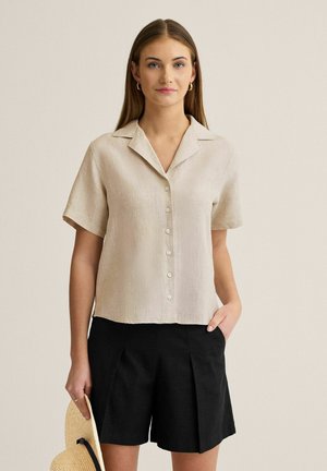 Kvinde iført beige kortærmet skjorte med knapper, sorte plisserede shorts, holder en stråhat med sort bånd, står mod en ensfarvet baggrund.