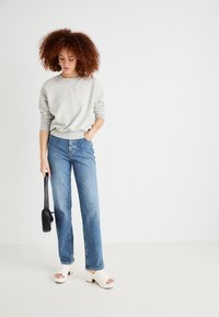Sweatshirt gris, coupe ample, col rond. Jean taille haute bleu clair, jambe droite. Sabots blancs ouverts à l'avant, sac à main noir. Textures lisses.