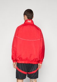 Rød vindjakke med elastiske manchetter og nederste kant, med en hvid accentlinje over ryggen og "Bulls" broderet på kraven.
