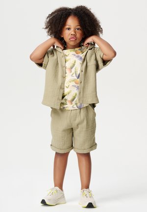 Ensemble de chemise à manches courtes texturée couleur marron clair et de shorts, avec un t-shirt à motif floral en dessous. Baskets légères avec des accents jaunes.