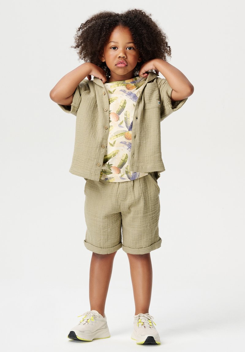 Ensemble de chemise à manches courtes texturée couleur marron clair et de shorts, avec un t-shirt à motif floral en dessous. Baskets légères avec des accents jaunes.