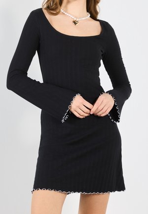 Kobieta w czarnej mini sukience z długim rękawem z białymi falbankami na brzegach oraz z perłowym naszyjnikiem z złotym zawieszką w kształcie serca.