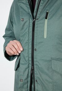 Giacca verde a zigzag con zip frontale, bottoni a pressione e una piccola tasca con zip. Il tessuto sembra testurizzato e resistente.