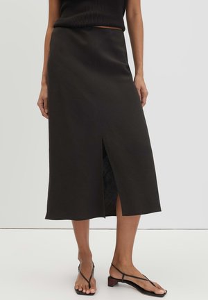 Woman wearing a black midi skirt with a front slit paired with black strappy low-heeled sandals, standing against a plain light background.