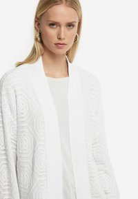 Vit stickad cardigan med ett geometriskt mönster i öppet arbete, med en ribbad krage och långa ärmar. Kraftig textur, lättviktsdesign.