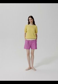 Žena stojaca s rukami v kapsách, oblečená v žltom svetri s krátkym rukávom, ružových krátkych nohaviciach a priehľadných slip-on topánkach na bielom pozadí.