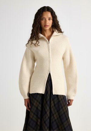 Donna con capelli ricci indossa un maglione crema a coste con zip e una gonna scura a quadri, in piedi davanti a uno sfondo bianco uniforme.