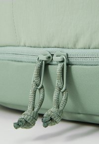 Bolsa acolchada de color verde claro con una superficie de tejido suave, que presenta un cierre de doble cremallera y tiradores trenzados decorativos.