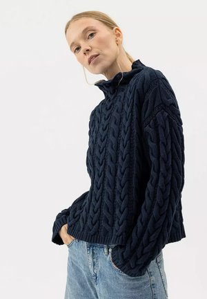 Femme portant un pull épais bleu marine à torsades avec un col montant, associé à un jean denim bleu clair, mains dans les poches.