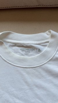 Vit kortärmad t-shirt med ribbad rund halsringning. Etiketten säger "Abercrombie & Fitch", storlek M, "Premium Essentials" och "Tillverkad i Vietnam."
