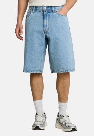Mann trägt hellblaue Jeansshorts, weiße Socken und weiße und schwarze Sneakers, steht mit einer Hand in der Tasche.