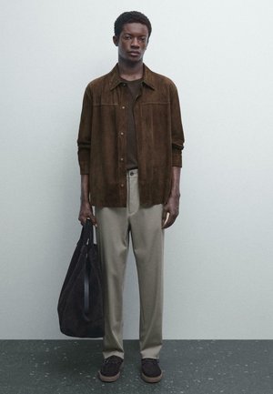Young man wearing a brown suede shirt, dark t-shirt, beige pants, black shoes, holding a large black tote bag, standing on a speckled floor.