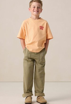 Smiling boy wearing peach "Venice Beach" T-shirt, olive green rolled-up pants, and tan slip-on shoes, standing with hands in pockets.