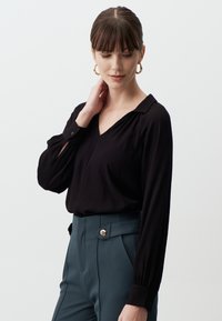 Blouse noire à manches longues avec un col en V et des épaules froncées, associée à un pantalon taille haute teal avec un accent de bouton doré.